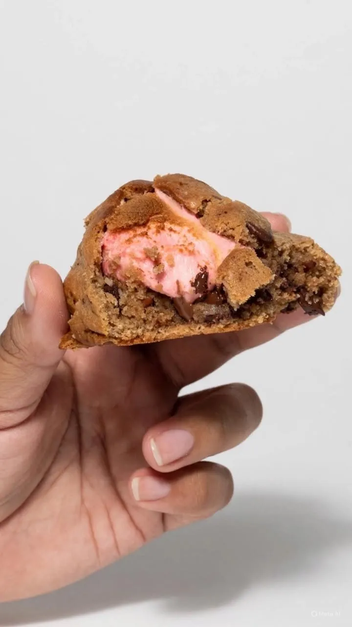 Mano sosteniendo una galleta con chispas de chocolate partida a la mitad, rellena de crema rosa