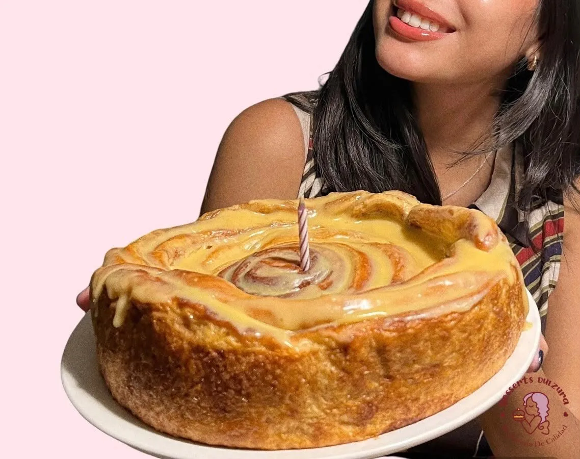 Rol de canela gigante con glaseado y una vela de cumpleaños, sostenido por una persona sonriente